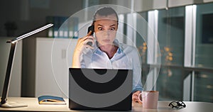 Angry businesswoman talking on smartphone while working at laptop in dark office