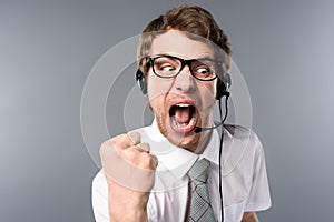 Angry call center operator in headset and glasses yelling and showing fist on grey background.