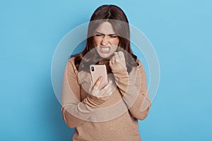 Angry casual woman using smart phone isolated on blue background, frowning face and clench fist with anger, looks at display,