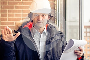 An angry disgruntled builder worker in a helmet with project drawings plans in his one hand and mobile phone in another