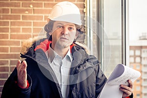 An angry disgruntled builder worker in a helmet with project drawings plans in his one hand and mobile phone in another