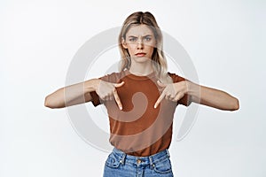 Angry and displeased young woman pointing fingers down, frowning upset, showing something disappointing, white