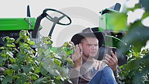 Angry farmer talking on the phone sitting in front of a green tractor in the field, problems at work