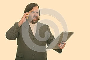 Angry fat Caucasian businessman talking on mobile phone while holding on clipboard