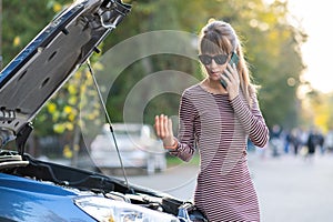 Angry female driver talking on cell phone with assistance service worker standing near a broken car with popped up hood on road