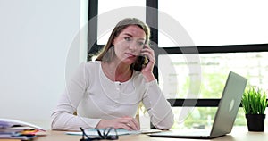 Angry female employee argues talking on mobile phone while sitting at desk at workplace in office