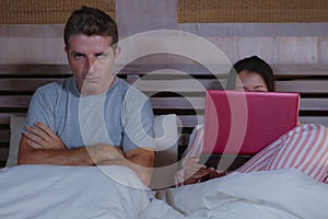 Angry and frustrated husband moody in bed ignored by his workaholic wife or internet social media addict girlfriend using laptop