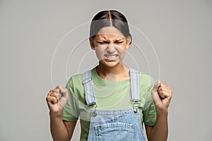 Angry frustrated teen girl clenched fists has evil frown face with closed eyes on grey background.