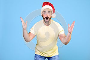 Angry and furious man in a Christmas hat on a blue background. He throws up his hands in rage and screams.