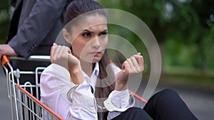 An angry girl in handcuffs sits in a shopping cart in business clothes. Boss man took the girl outdoors handcuffed to