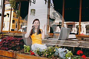 Angry girl in outdoors street cafe sitting at table with laptop pc computer, talking on mobile phone, screaming and