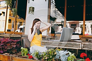 Angry girl in outdoors street cafe sitting at table with laptop pc computer, talking on mobile phone, screaming and