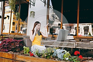 Angry girl in outdoors street cafe sitting at table with laptop pc computer, talking on mobile phone, screaming and