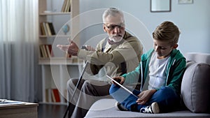 Angry grandfather scolding boy for playing video game on tablet, generation gap