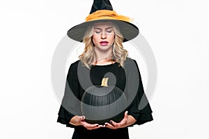 Angry Halloween Witch holding large black pumpkin, looking down on it. Beautiful young woman in witches hat and costume.