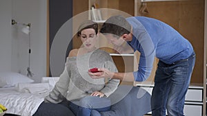 Angry husband showing messages in smartphone to guilty cheating wife sitting on bed at home. Furious Caucasian man