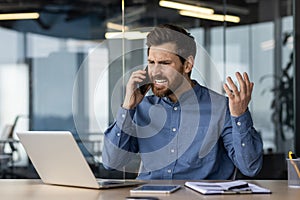 Angry and irritated young businessman sitting in the office at the desk and talking on the phone while waving his hands
