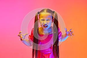 Angry little girl in festive dress with ponytails posing over pink neon background. Concept of childhood, emotions, kids