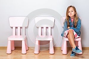 Angry little girl sitting on a chair against a gray wall. Empty chairs next to the child