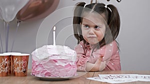 An angry little girl is sitting at the festive table next to the birthday cake. Something went wrong.