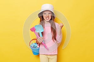 Angry little girl wearing casual clothing and panama holding sandbox toys and bucket isolated over yellow background standing with