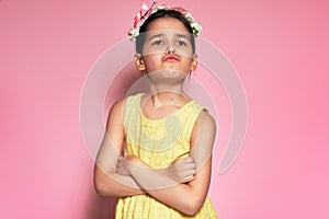 An angry little girl wearing yellow dress standing with crossed arms posing over pink background. Displeased preschooler child