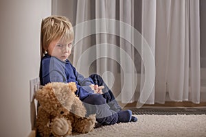 Angry little toddler child, blond boy, sitting in corner with teddy bear, punished