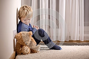 Angry little toddler child, blond boy, sitting in corner with teddy punished for mischief photo