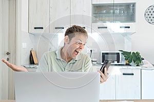 Angry mad man screaming, yelling and arguing talking on smartphone while working on laptop online at home. Upset male