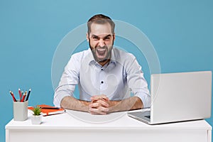 Angry mad young bearded man in light shirt sit work at desk with pc laptop isolated on pastel blue background