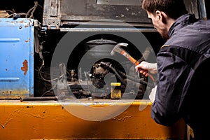 An angry male mechanic is repairing the engine of an old truck with an open hood hitting it with a hammer