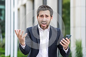 An angry man, a businessman, shouts at the camera, holds a mobile phone in his hands, spreads his hands in displeasure