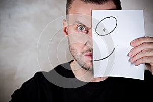 Angry man covers half his face with a piece of paper with a painted smile