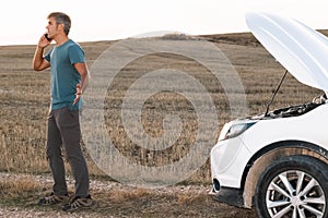 An angry man in the middle of the field calling the insurance of his broken down car for assistance at sunset