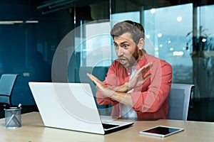 An angry man in the office talking on a video call from a laptop. Gestures with his hands, denies