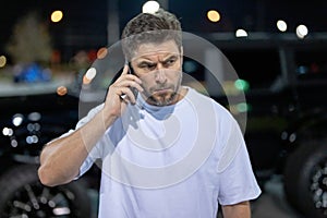 Angry man talking on phone near car on night urban street. Dangerous aggressive man talking on phone with serious face