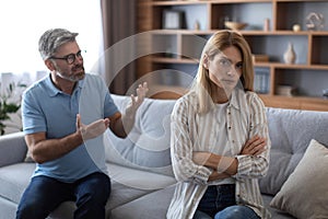 Angry mature caucasian man scolds to female on couch, quarreling in living room interior