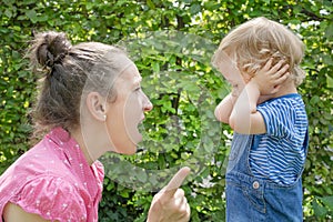 Angry mother shout at her naughty son in the park while the little child cover his ears avoiding mad mom screaming at him. Parent