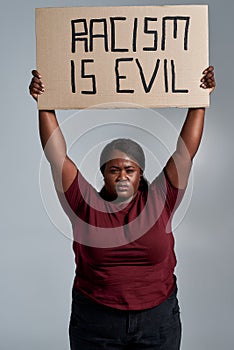 Angry plus size young african american woman in casual clothes looking at camera, holding Racism is evil banner above