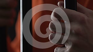 Angry prisoner in orange jumpsuit holding cell bars, serial killer hands closeup