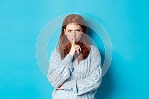Angry redhead girl hushing at you, show taboo gesture, forbid to speak, standing over blue background in sweater