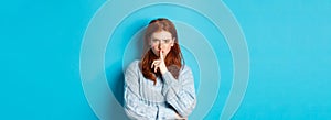 Angry redhead girl hushing at you, show taboo gesture, forbid to speak, standing over blue background in sweater