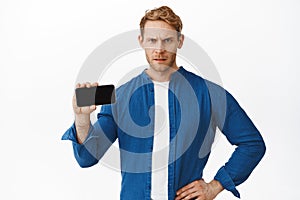 Angry redhead man shows horizontal smartphone screen and frowning judgemental, showing something bad and disappointing