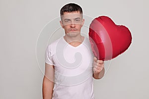 Angry sad unhappy young male carries heart shaped balloon standing isolated over gray background waiting his girlfriend for date