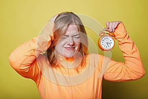 Angry sad woman holding her head with one hand and orange big clock in other over yellow background. Depression deadline concept