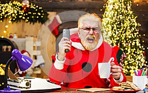 Angry Santa Claus sitting in armchair watching Christmas movie and drinks coffee. New Year cinema.