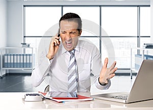 Angry senior businessman in stress working and talking on mobile phone at computer desk
