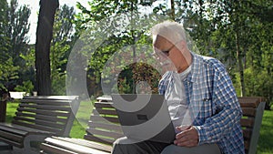 Angry senior man talks in skype, sitting on the bench in the garden. A pensioner is holding a laptop on his lap. He