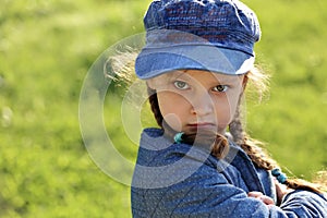 Angry serious kid girl in blue hat grimacing on summer green grass background. Closeup portrait