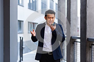 Angry and serious Successful Asian businessman explains information to employees using phone, speaks near office outside
